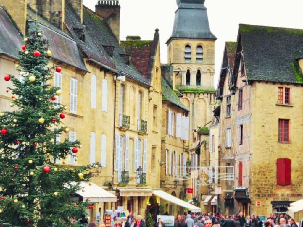 This image features a festive street scene in what appears to be the medieval town of Sarlat, France, during the holiday season. The street is lined with historic stone buildings, and a large Christmas tree decorated with red and gold ornaments stands prominently in the foreground. Crowds of people are gathered, likely exploring the Christmas market, while the steeple of a historic building looms in the background, adding to the quaint charm of the setting.