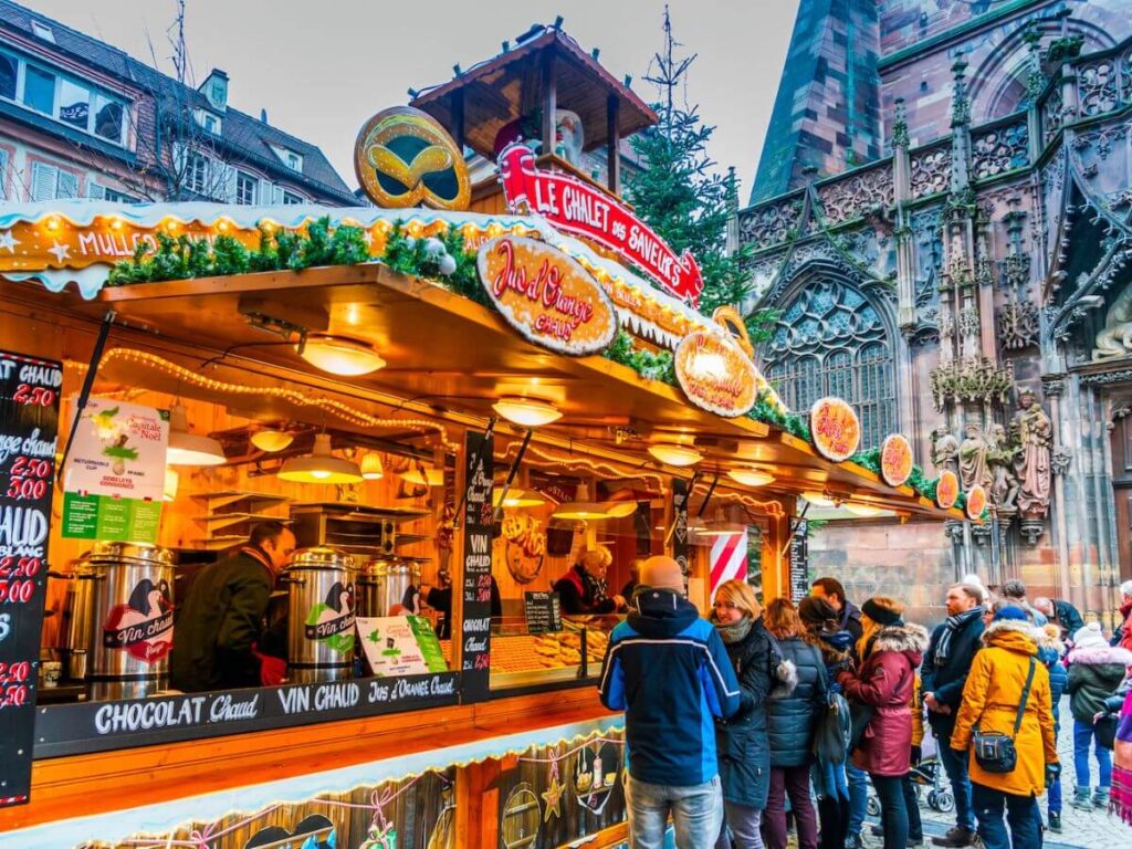 A Christmas Market Stall selling food and drink in front of a cathedral with people walking around during the day