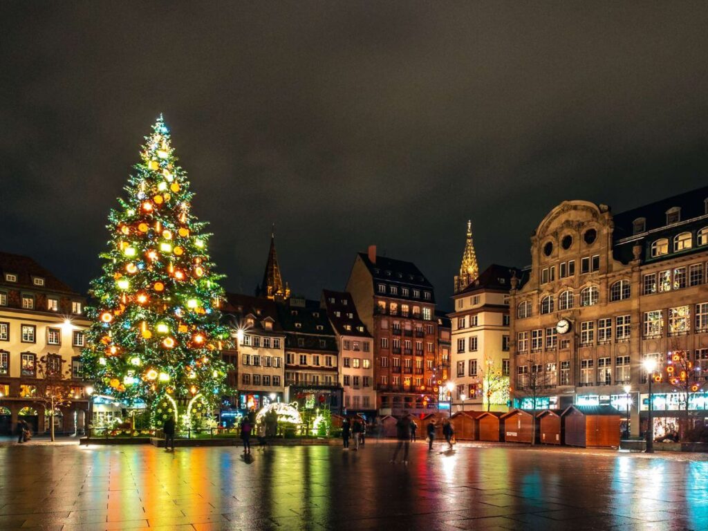 A tall Xmas Tree lit at night and reflected in the square in front of it with lit buildings behind