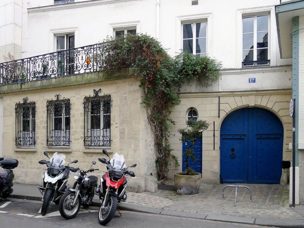 Two motorcycles sat on the road outside a building with a lattice balcony and an arched blue garage door