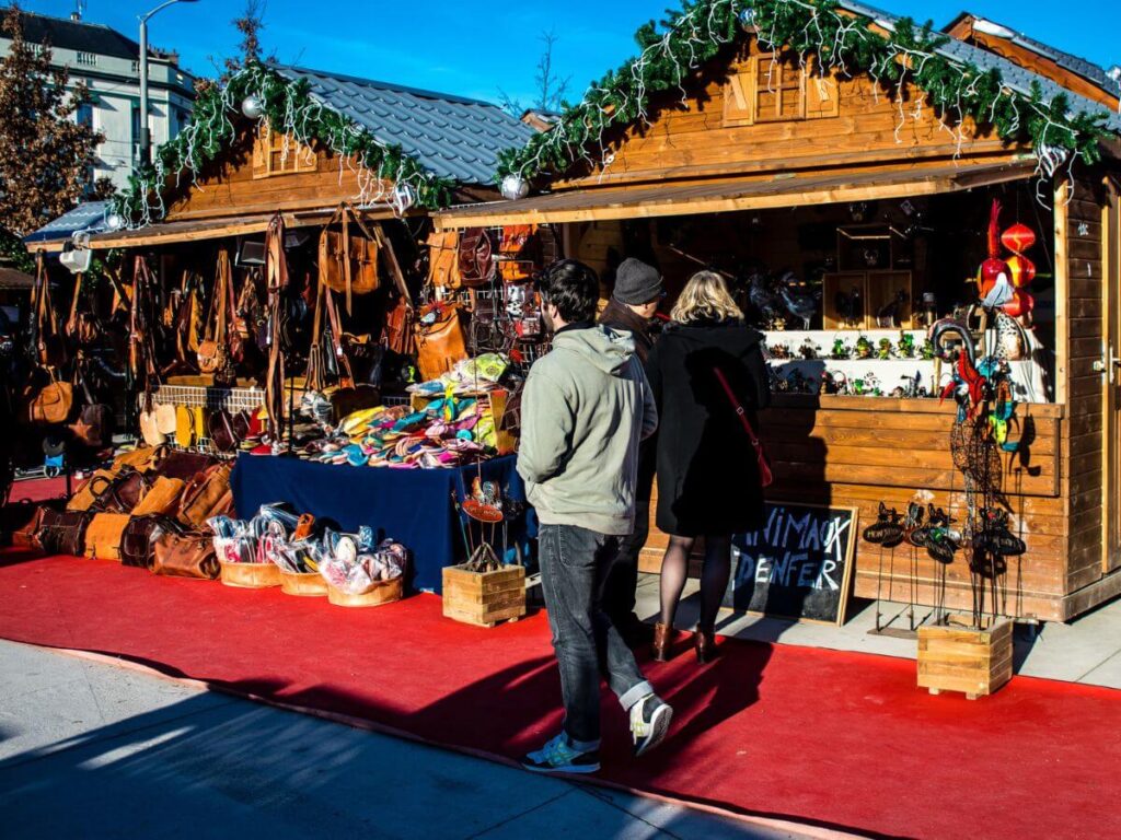 Wooden chalets at the Reims Christmas Market with a red carpet in front and people looking at the stalls with a blue sky overhead
