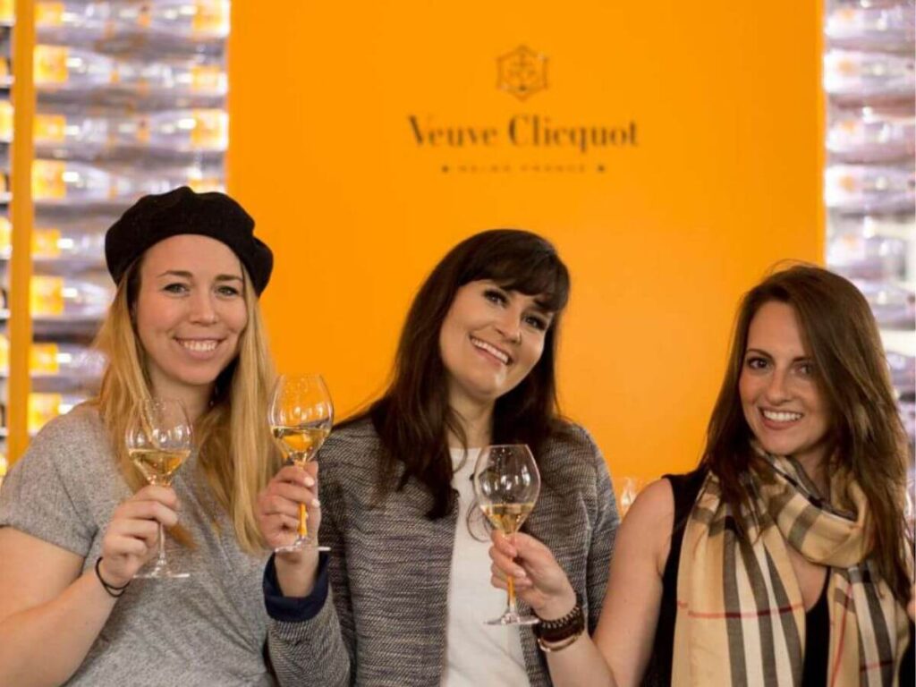 3 Women holding champagne glasses stood in front of an orange veuve cliquot sign at the winery