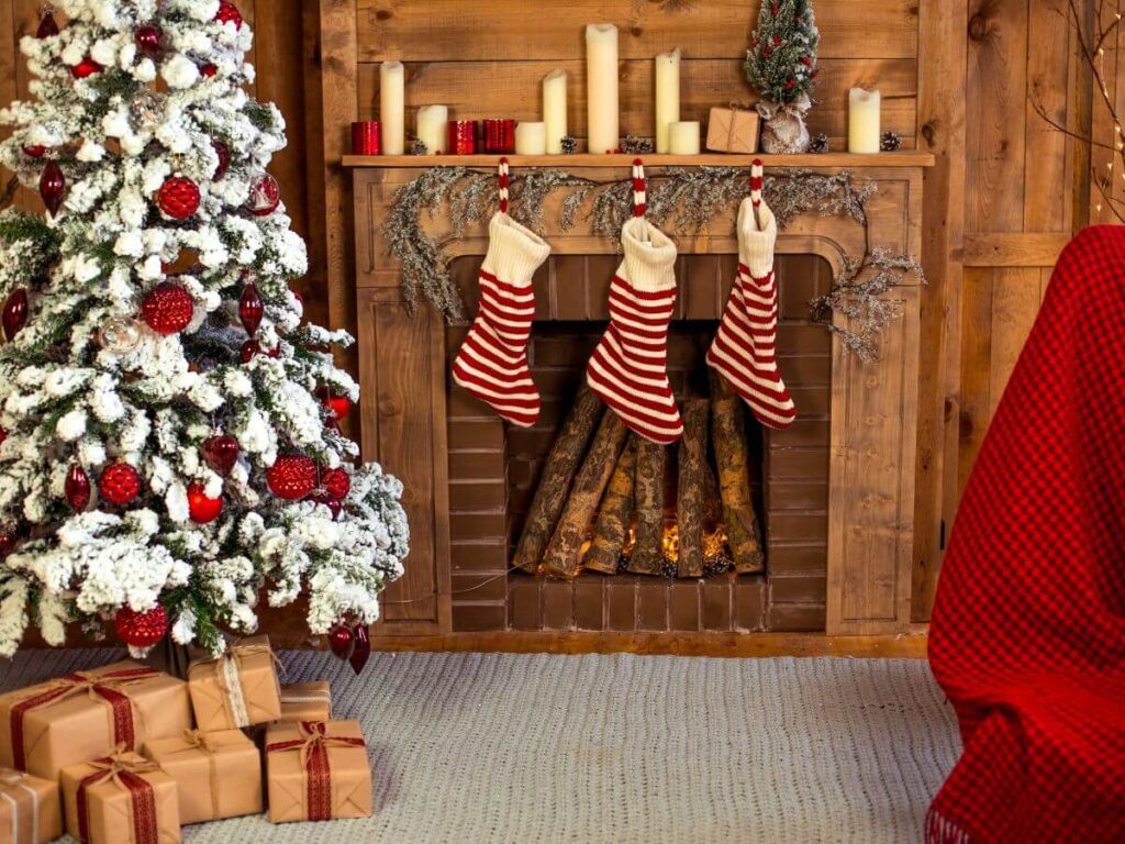 A Christmas tree in a lounge by a fire covered in fake snow with presents wrapped in brown paper underneath. There are three red and white striped stockings hanging from the firel place with candles on the mantlepiece