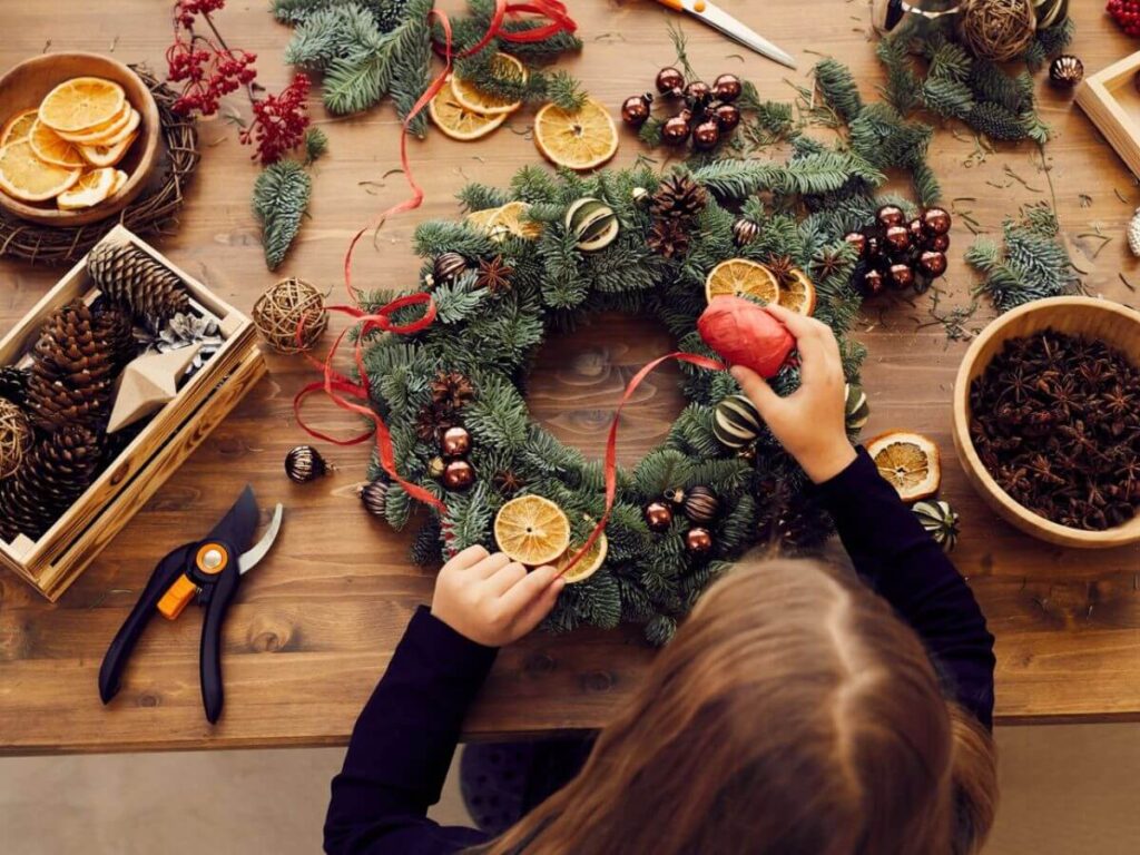 A young girl sat at a wooden table making a Christmas wreath with red ribbon, and fruit using scissors