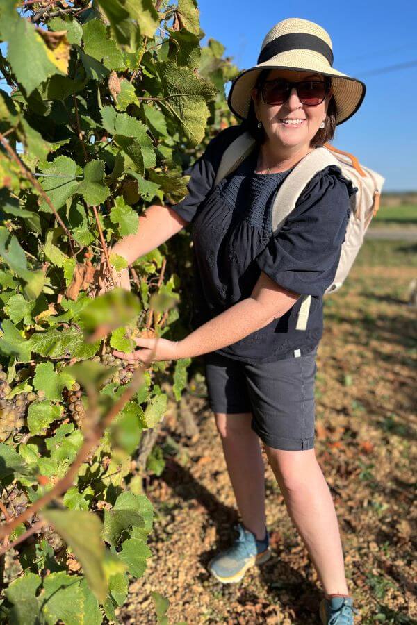 A woman in blue shorts and a blue top with a sun hat and sunglasses holding some grapes on a vine