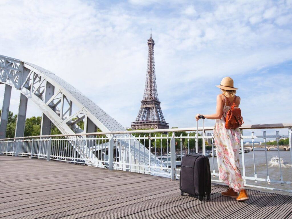 A woman with a suitcase wearing a summer dress, backpack and hat walking towards the Eiffel Tower