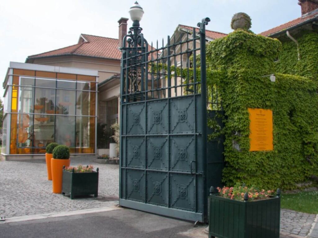 The outside of the Veuve Cliquot Champagne House wih a big grey wrought iron gate and orange sign with some flower baskets