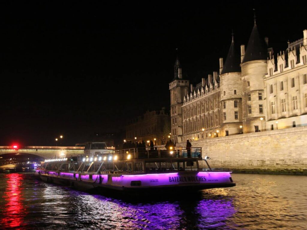 A cruise boat on the Seine River at night with purple lights and a cream building on the right hand side of the river