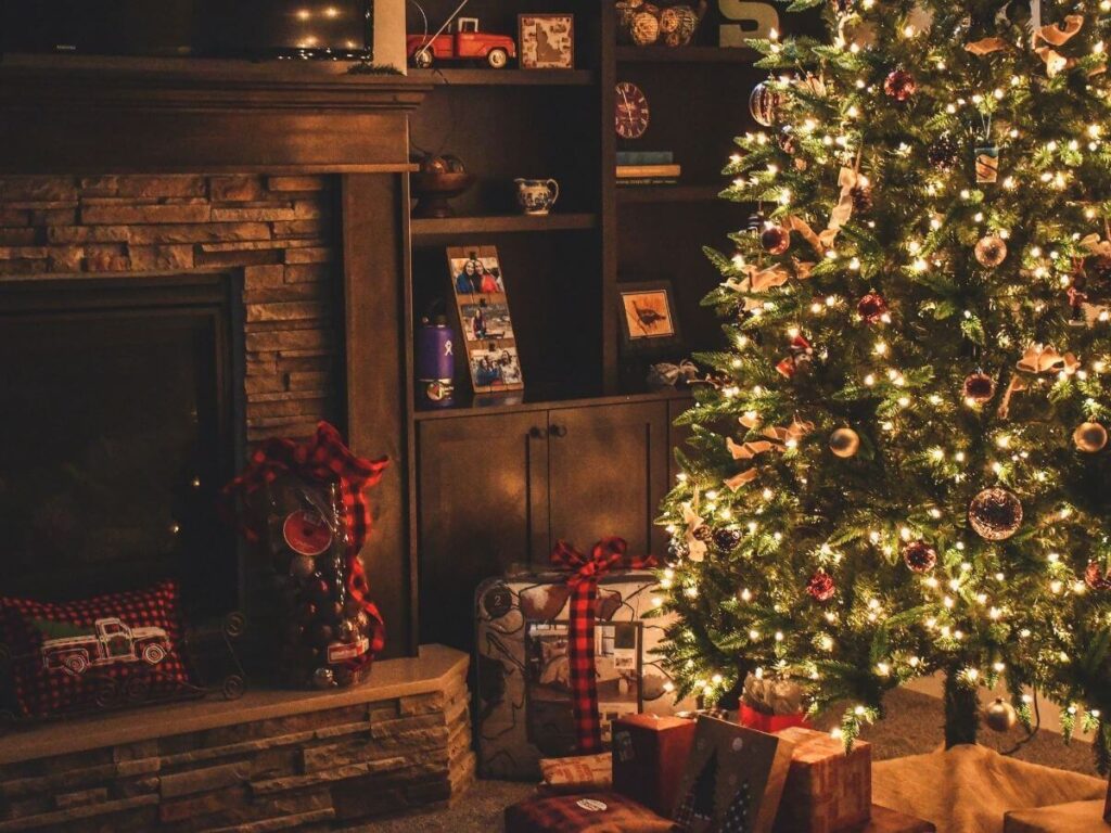 A Christmas Tree at night all ilt up in a lounge room with a fireplace on the left. There are boxes wrpaaed in brown paper under the tree.