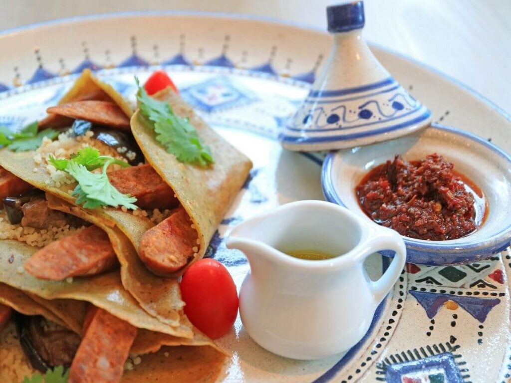 A plate with savoury crepes filled with sausages, lettuce and tomatoes with a bowl of minced meat beside it and a white jug with some sauce all sat on a blue and white tray