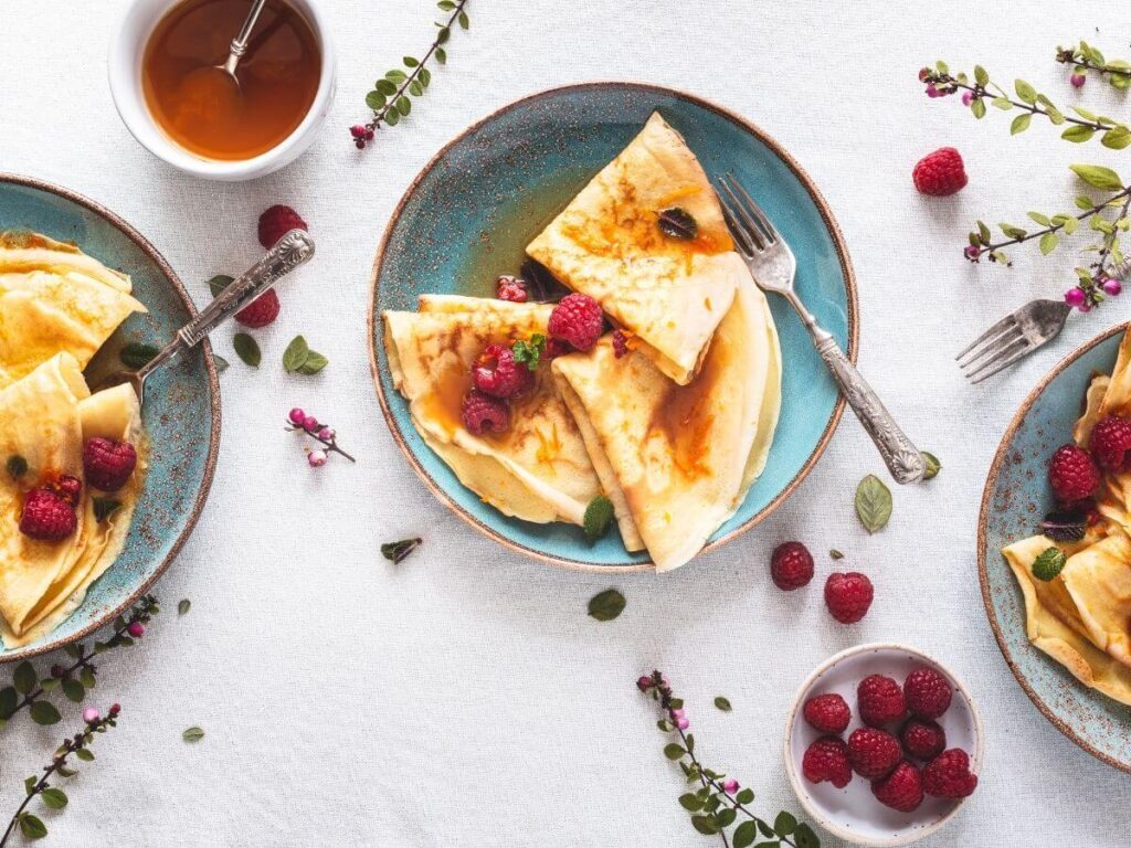 Three blue plates with crepes all with raspberries on top and a bowl of syrup on the side