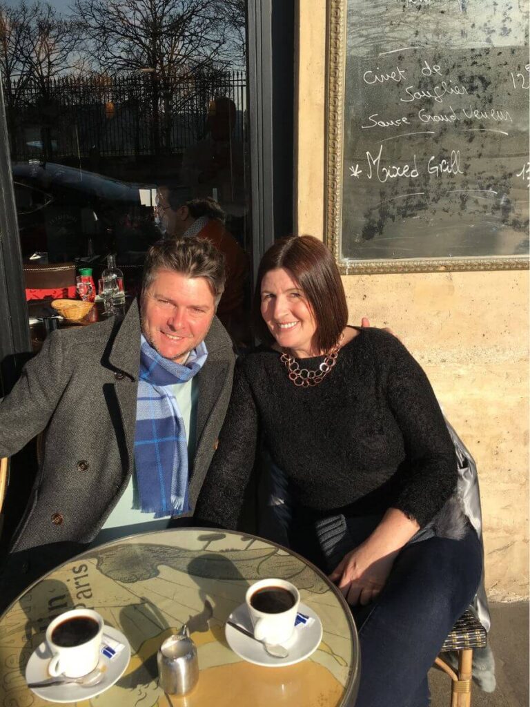 Two people sitting outside a French cafe drinking coffee in France. The man has a gray winter coat and blue scargf and the woman is wearing a black jump and it's sunny