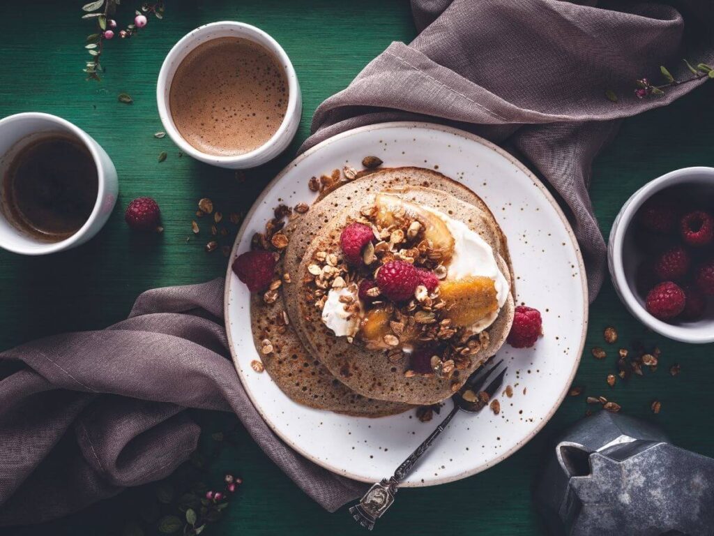 A plate with buckwheat cre[es with topping of nuts, cream and raspberries sat on a piece of brown material with a cup of coffee by the side.