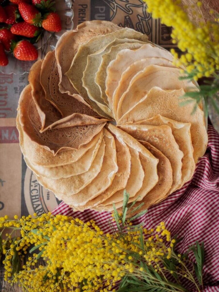 A plate of crepes with some yellow flowers sat either side and sitting on a pink cloth with some strawberries