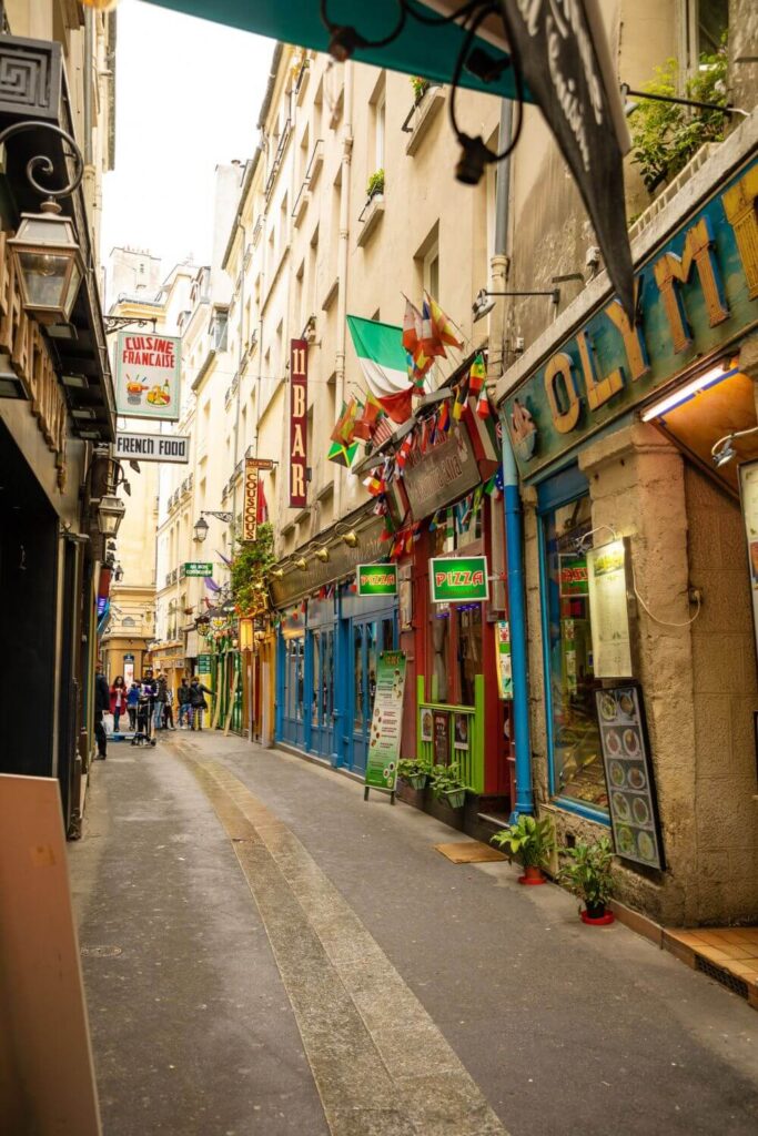 An empty street in the Latin Quarter of Paris with colourful doorways and shop fronts. There are many country flags hanging out ot eh windows as well