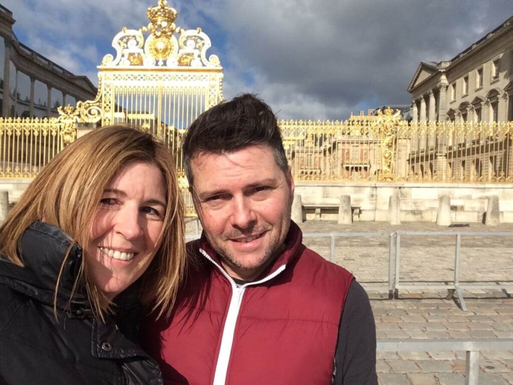 A man and a women smiling at the camera stood outside the gold gates of the palace of Versailles in France