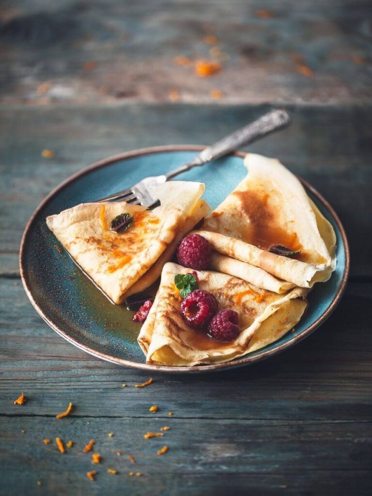 A plate with several small crepes folded into traingles with raspberries on top sat on a blue plate wiht a fork