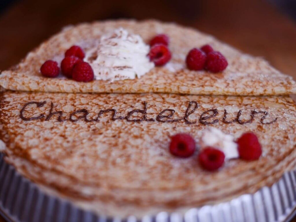 A giant crepe on a plate with the word Chandeleur written in chocolate on it with some raspberries and ice cream on top