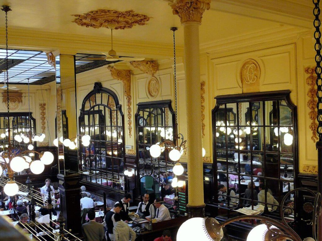 The image displays the bustling interior of a grand Parisian bouillon restaurant, viewed from an upper level. The décor features elegant, gilded columns and ornate plasterwork with floral motifs adorning the ceiling. Large mirrors with black frames enhance the sense of space, reflecting the glow of spherical pendant lights that hang throughout the venue. Waitstaff in traditional attire navigate between the tables, attending to patrons. The atmosphere is lively, with a sense of historic charm highlighted by the classical architectural elements and the warm, ambient lighting.