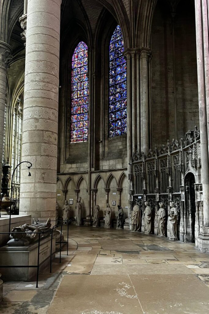 This image captures the interior of Rouen Cathedral with a focus on the high Gothic arches and stained glass windows that cast colorful light into the space. Stone statues line the nave, leading the eye towards the altar in the distance. The cathedral's grandeur is evident in the height of its vaulted ceilings and the elegance of its supporting columns.