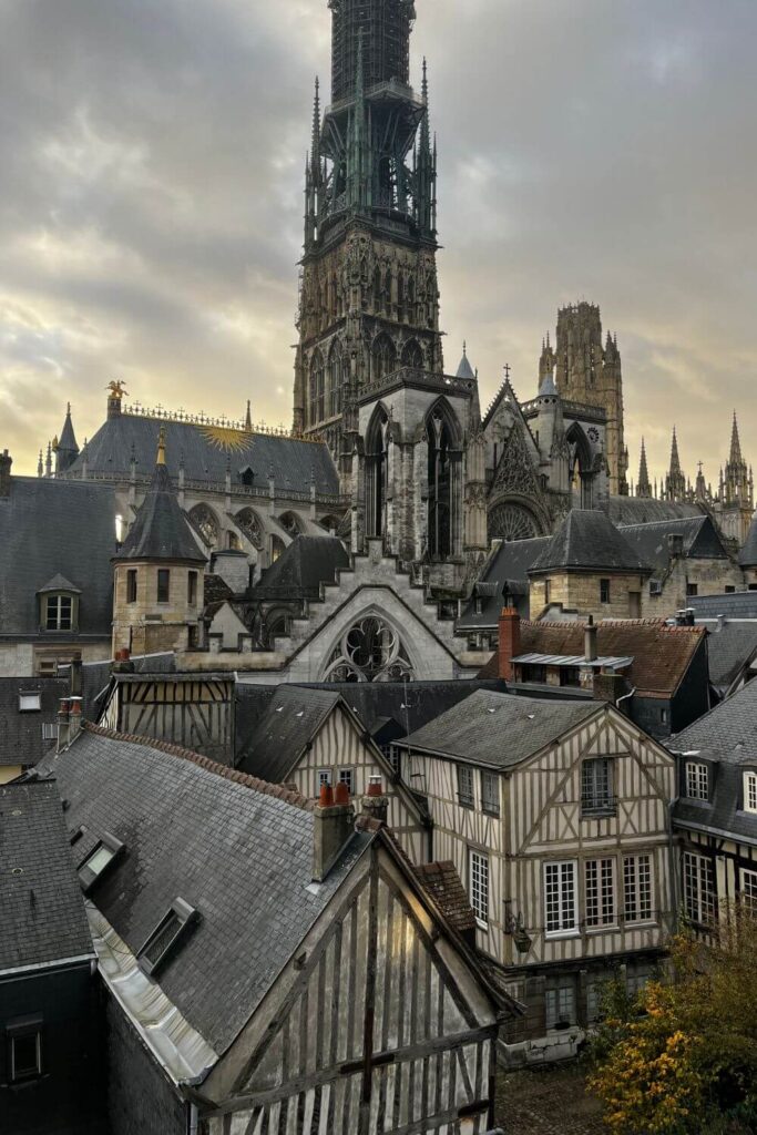 A stunning elevated view of Rouen, showcasing the intricate Gothic architecture of Rouen Cathedral, which dominates the skyline with its elaborate spire. The cathedral's complex façade and flying buttresses are visible against a moody sky, suggesting either early morning or late afternoon light. In the foreground, traditional half-timbered houses with steeply pitched roofs, typical of Normandy's architectural style, cluster tightly together. The muted tones of the buildings, consisting of browns, beiges, and greys, complement the historic and somber atmosphere of the scene. The overall image captures the essence of Rouen's historical charm, blending imposing religious structures with the quaint, cozy feel of the city's old residential buildings.