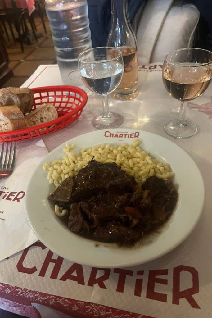 A plate of Boeuf Bourguignon served with coquillettes pasta at Bouillon Chartier. The meal is accompanied by glasses of water and wine, a bottle of water, and a basket of bread on a table covered with Chartier branded placemats.
