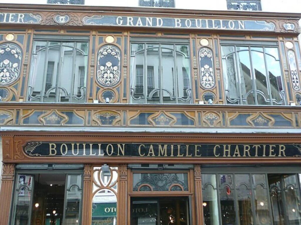 The photograph showcases the façade of the Grand Bouillon Camille Chartier, a renowned Parisian restaurant. The exterior is ornately decorated with Art Nouveau motifs, featuring elegant gold and blue patterns on a dark wooden backdrop. The upper part of the façade displays large windows with intricate ironwork, offering a glimpse into the two-story interior. The restaurant's name is prominently displayed in bold, capital letters above the entrance, beneath the phrase "Grand Bouillon," inviting patrons to experience a piece of Paris's culinary history.