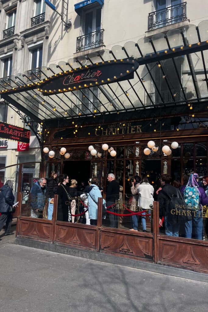 The exterior of Bouillon Chartier Montparnasse with a crowd of people waiting outside. The entrance features a classic French café design with a sign reading 'Chartier' illuminated by yellow lights.