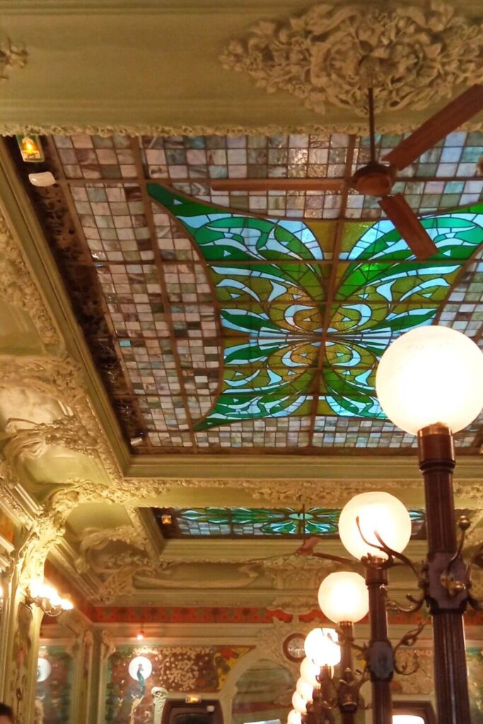 The image captures the detailed ceiling of a Parisian bouillon restaurant, highlighted by a stained glass panel with a prominent green and yellow pattern. Surrounding the glasswork is an ornate ceiling with plaster reliefs and intricate moldings, featuring floral and scroll motifs. The ambiance is enhanced by warm lighting from spherical, frosted glass lamps mounted on elegant bronze fixtures. The ceiling's rich decoration and the interplay of light and color contribute to the establishment's historic and luxurious atmosphere.