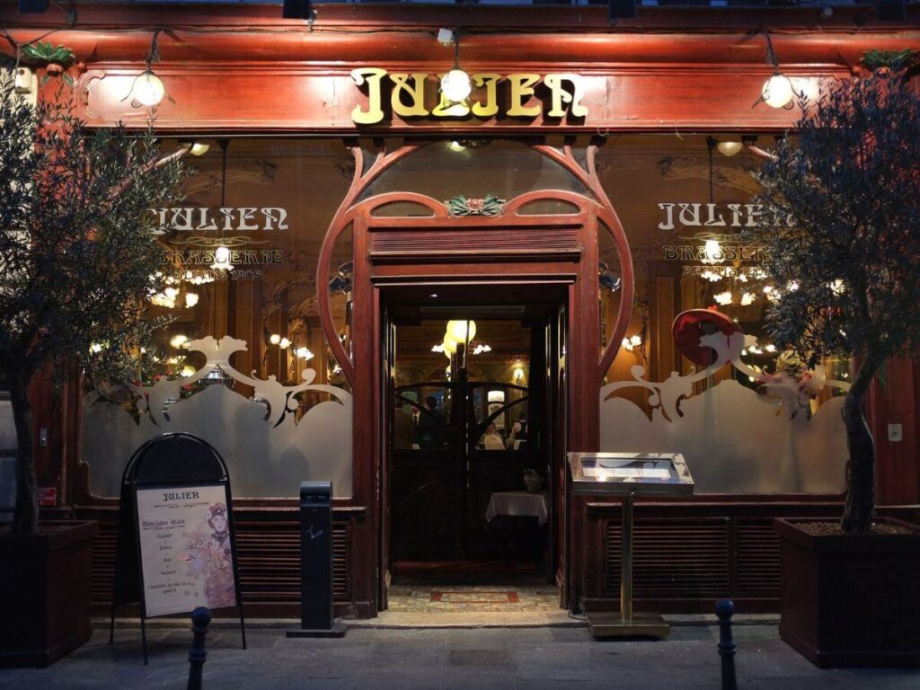 The exterior of Bouillon Julien, a classic Parisian brasserie, is shown in the evening. The façade is warm and inviting, with its rich wooden trim and ornate Art Nouveau style etched windows that display the restaurant's name. Frosted glass panes feature elegant white designs, complementing the graceful, curving woodwork. Small trees in wooden planters flank the entrance, and a traditional menu board stands to the side, detailing the day's offerings. The lighting, signage, and historical architecture convey the timeless appeal of this dining establishment, which has been serving patrons since 1903.