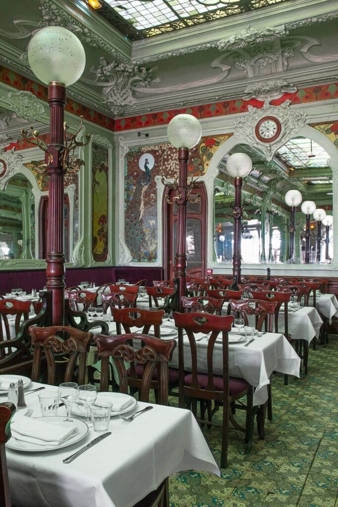 The interior of Bouillon Julien in Paris is elegantly displayed, with Art Nouveau style clearly influencing the décor. Tall wooden columns with ornate tops support frosted globe lamps, casting a soft light over the dining area. The ceiling boasts intricate mouldings and an elaborate skylight, while the walls are adorned with mosaic tiles and swirling patterns. Rich, dark wooden chairs with curved backrests are neatly arranged around white-clothed tables set with plates, cutlery, and glasses. The patterned floor ties the room together, reflecting a bygone era of luxurious design and fine dining.