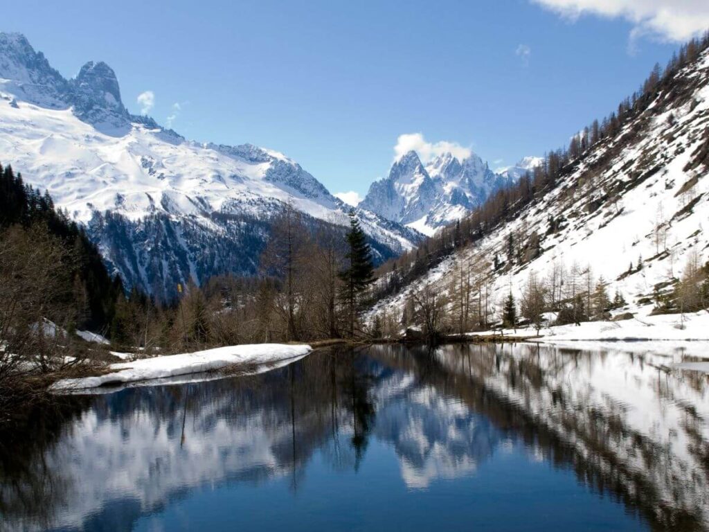 A serene winter landscape showcasing a tranquil lake reflecting the surrounding snowy peaks and bare trees under a clear blue sky, in a valley suggestive of the Chamonix region, renowned for its alpine beauty and winter sports appeal.