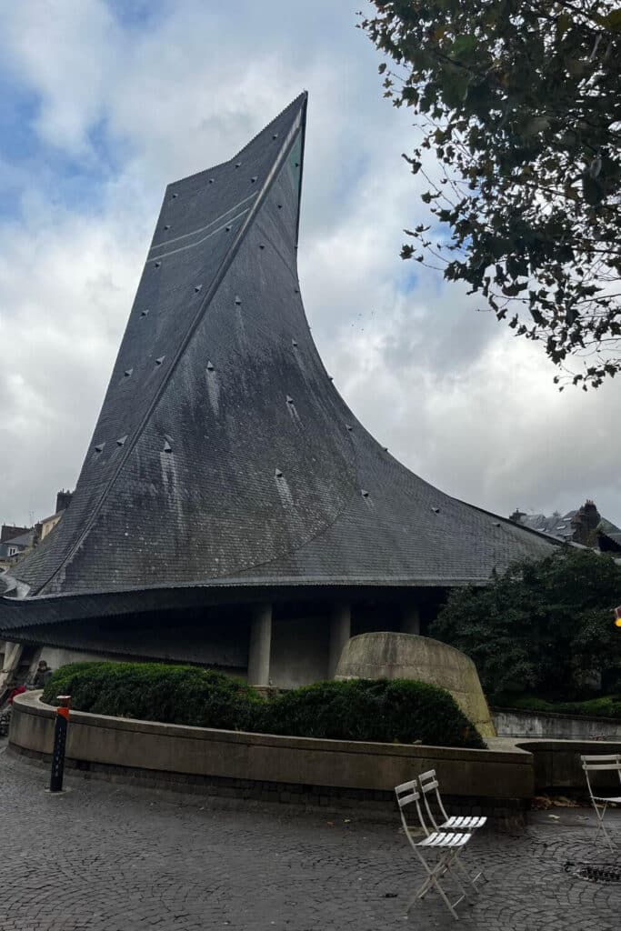 The picture captures the distinctive, sloping roof of the Church of Saint Joan of Arc in Rouen, rising sharply against a cloudy sky. The church's unique silhouette, reminiscent of an upturned ship's hull, is a modern interpretation amidst the city's historic architecture, symbolizing both the flames that consumed Joan of Arc and a gathering of the faithful.