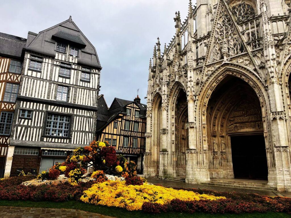 This image features the Church of Saint-Maclou, a masterpiece of the Flamboyant Gothic style, situated in Rouen, France. In the foreground, vibrant flowerbeds in shades of yellow and maroon create a striking contrast against the gray cobblestone plaza. The background displays the church's elaborate façade with its finely carved stone arches and towering spires. Adjacent to the church are traditional half-timbered houses, typical of Normandy's architectural heritage.