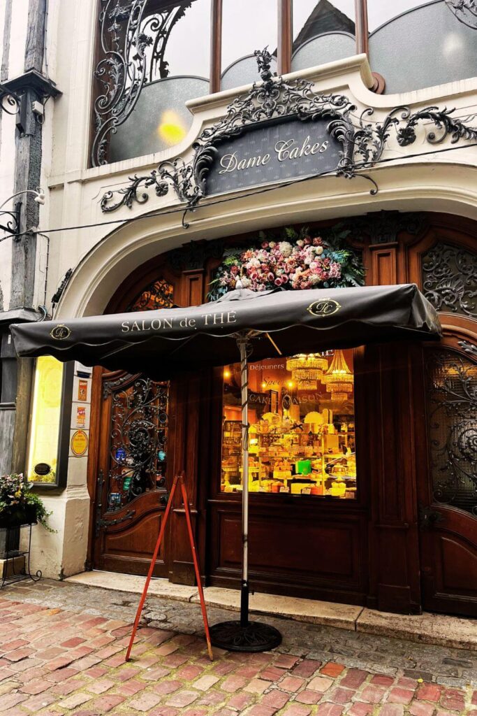 The storefront of 'Dame Cakes,' a tea room in Rouen, France. The sign above the entrance features elegant wrought-iron work and sophisticated script against a black background. Beneath a decorative awning labeled "SALON de THÉ" is a beautiful wooden door flanked by large windows showcasing an inviting display of cakes and pastries. The door is adorned with intricate carvings, and a lush flower arrangement sits above the awning, adding a splash of color to the façade. A sandwich board sign stands on the cobblestone pavement in front, hinting at a cozy spot where visitors can enjoy tea and homemade sweets. The architecture and décor suggest a blend of traditional craftsmanship with a touch of luxury, inviting passersby to step in and savor the delicacies within.