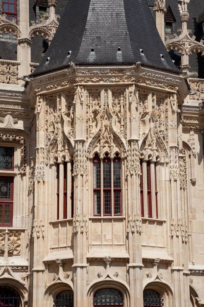 The photo provides a close-up of the Palais de Justice's detailed stonework in Rouen. The gothic flamboyant style is evident in the ornate tracery and the towering windows with pointed arches. The façade is a light stone color, with each architectural element sharply defined, from the delicate finials to the statues nestled in niches, all set against a clear blue sky.