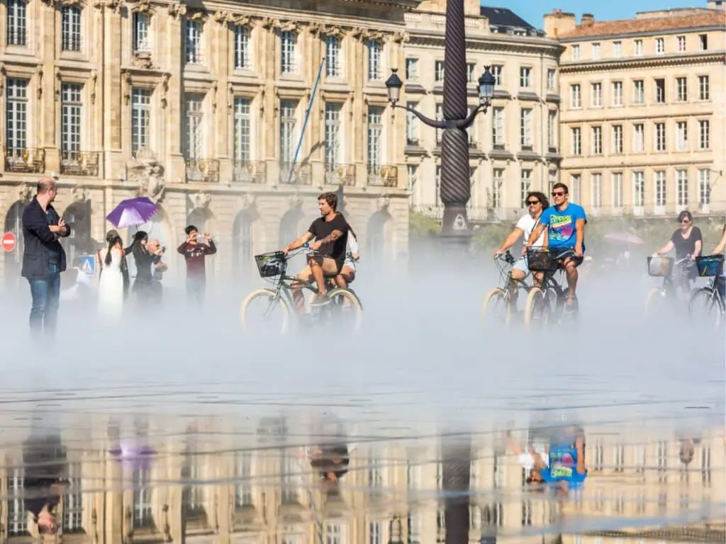 People riding bikes through the water mirror in Bordeaux on a hot day with cream buildings in the background.