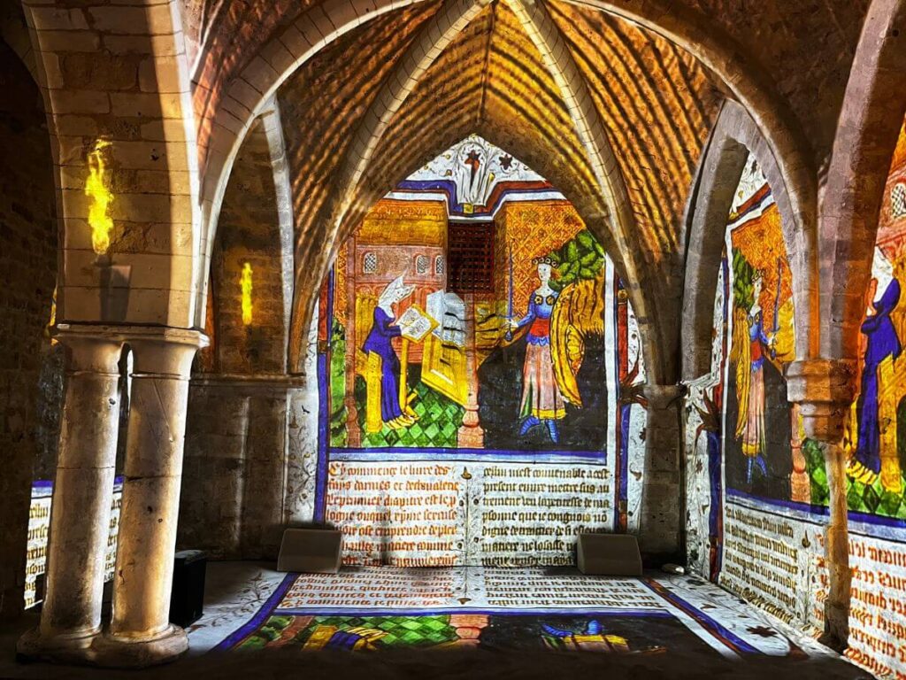 The image captures a vibrant stained-glass window inside the Historial Jeanne d'Arc in Rouen. The window depicts colorful scenes with figures in medieval attire, casting vivid reflections onto the ancient stone floor and vaulted Gothic arches of the interior. The warm illumination contrasts with the dark, rustic stonework, creating a dramatic and immersive atmosphere.