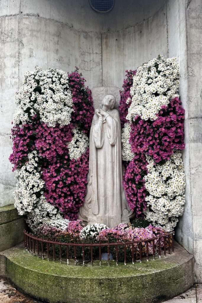 The photograph shows a stone statue of Joan of Arc in prayer, flanked by vibrant floral arrangements of white and purple chrysanthemums. The statue is nestled within a semi-circular alcove, and a protective railing encircles the display. The presence of the statue offers a quiet space of remembrance amidst the bustling city environment.