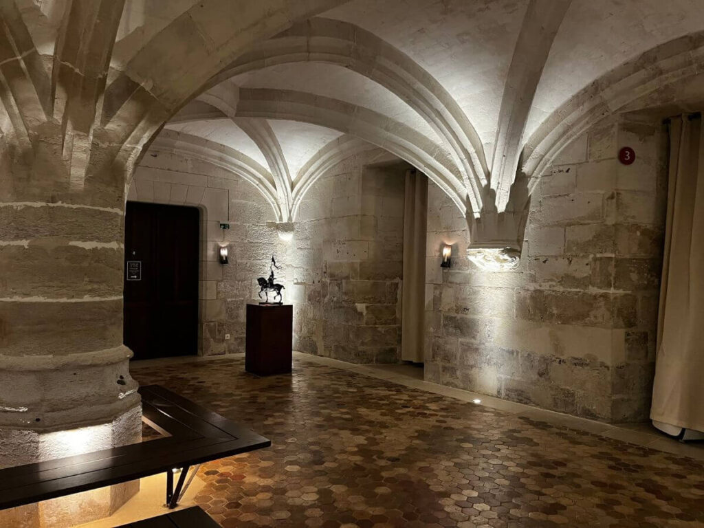 A photograph of a dimly lit, vaulted stone room resembling a crypt, with patterned cobblestone flooring. A solitary bench sits in the center, and a small sculpture is displayed on a pedestal, adding an artistic focal point to the space. The room is likely part of a historical building, featuring medieval architecture that evokes a sense of solemnity and timelessness.