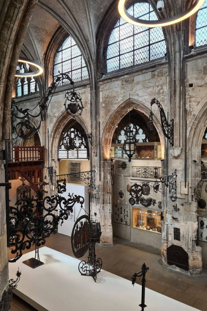 The interior of the Musée Le Secq des Tournelles in Rouen, housed in an old Gothic church, showcases an extensive collection of wrought ironwork. The exhibition is displayed against the backdrop of tall stained-glass windows and stone arches, with a variety of intricate metal pieces, from delicate to grand, suspended throughout the space, creating a unique contrast between the medieval architecture and the crafted metalworks.