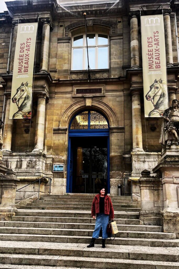 A woman stands on the steps of the Musée des Beaux-Arts in Rouen, France. The museum entrance displays classic architecture with a grand arched doorway and a large window above. Two vertical banners featuring artwork flank the entrance, announcing the museum's focus on fine arts. The stone façade and intricate decorative elements above the door give the building a dignified presence.