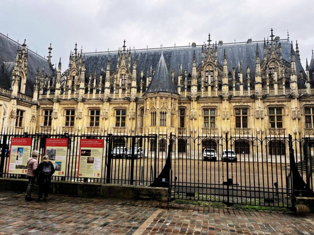 This image shows the Palais de Justice in Rouen, an impressive example of Gothic architecture. The building's façade is ornate with a series of gables adorned with intricate carvings and finials. A black wrought-iron fence runs along the front, behind which informational panels are mounted for visitors. Two people are examining the panels, providing a sense of scale against the grandeur of the historic courthouse.