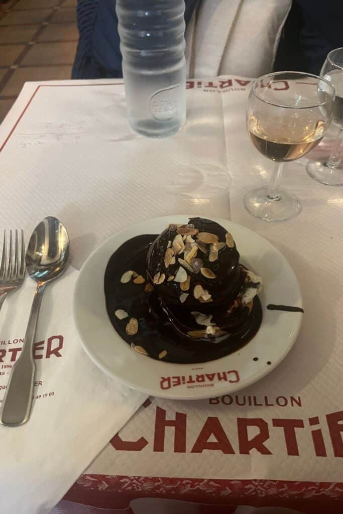 A plate of profiteroles topped with chocolate sauce and sprinkled with almond slices at Bouillon Chartier. The dessert is placed on a table with a spoon and fork, alongside a bottle of water and glasses of wine.