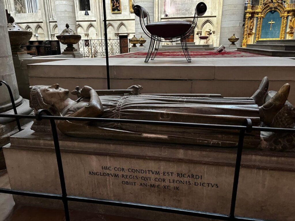 The image features a close-up of a recumbent statue believed to represent Richard the Lionheart, inside Rouen Cathedral. The effigy is highly detailed, with the figure depicted in repose, hands clasped in prayer. An inscription in Latin is visible on the base of the tomb, adding to the historical significance of the monument.