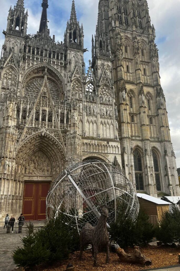 The photograph showcases the magnificent facade of Rouen Cathedral (Notre-Dame de Rouen) with its intricate Gothic architecture. The cathedral towers with its spires reaching towards the cloudy sky, and a detailed rose window is partially visible. In the foreground, a contemporary metal and wire sculpture of a deer enclosed in a spherical frame contrasts with the historic backdrop, creating a blend of old and new.