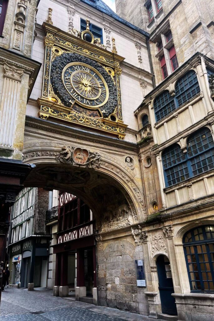 This image showcases a wider view of the Rue du Gros-Horloge in Rouen, with the Gros Horloge visible on the right. The intricate details of the arch and the clock are more apparent from this angle. The street is lined with traditional half-timbered buildings, and cobblestones pave the way. The historical architecture of the area provides a sense of Rouen's rich past, with modern elements subtly integrated, like the bollards at the street's edge.