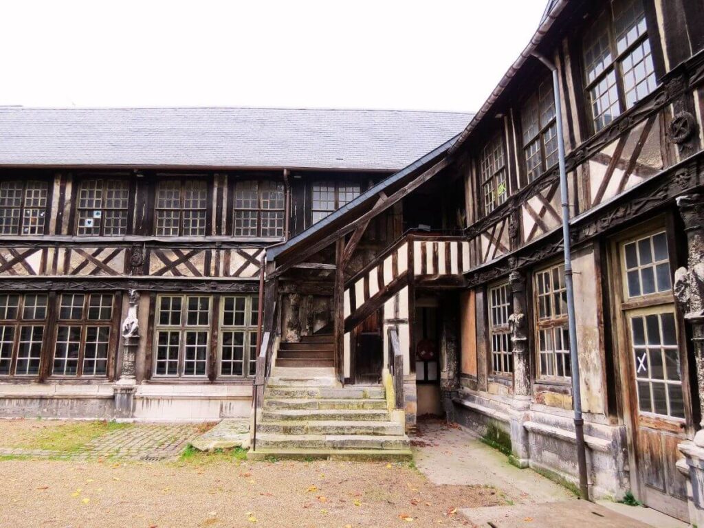 A view of the Saint-Maclou Ossuary in Rouen, an example of medieval half-timbered architecture. The building features exposed wood beams in a geometric pattern against white infill. A stone staircase leads to a wooden door, reflecting the historical function of the building associated with the rites of the dead.