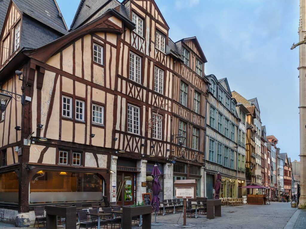 A quaint street lined with traditional half-timbered houses, characteristic of certain historical regions in France. The buildings are multi-story, with the upper floors often jutting out over the lower ones, displaying the typical wood and plaster exterior that forms a pattern of vertical, horizontal, and diagonal lines. The ground floors appear to be commercial spaces, some of which are cafes with outdoor seating comprising tables and chairs, now unoccupied. The street is cobblestoned, and despite the absence of people, the presence of open business signs suggests daytime. The sky is clear, hinting at good weather. This setting evokes a sense of stepping back in time and is typical of the architecture found in Rouen, a city in Normandy, France.