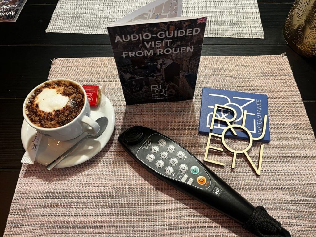 A tabletop scene from a visitor's perspective in Rouen. There is a cup of cappuccino with a heart-shaped design in the foam, sprinkled with chocolate, accompanied by a small packet of sugar. To the right, there's a stylish souvenir coaster with the word "ROUEN" in bold, decorative lettering. An audio guide device with buttons lies next to the coaster, indicating the availability of an audio tour. In the background, a folded brochure advertises an "Audio-Guided Visit from Rouen," available in multiple languages, including English, as denoted by the UK flag. This setting suggests a leisurely break during a touristic exploration of Rouen, combining the enjoyment of a local café with the cultural experience of the city.